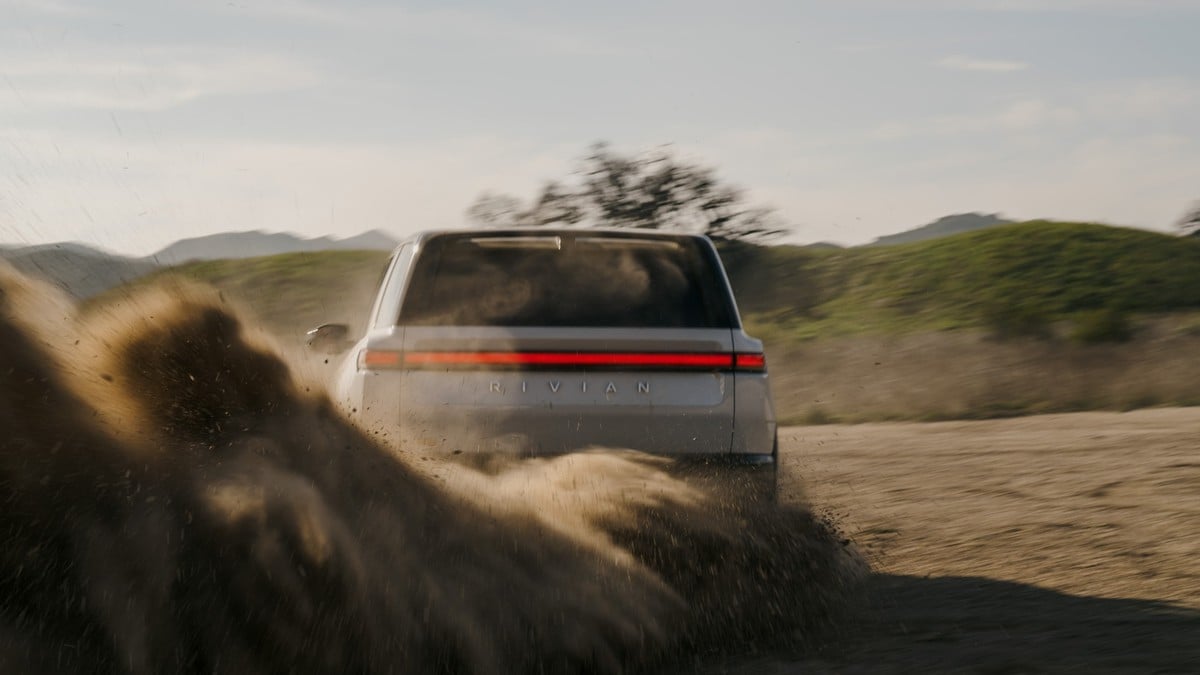 Rear view of a gray Rivian R2 electric SUV driving off-road and kicking up dust on a dirt trail in a hilly landscape.