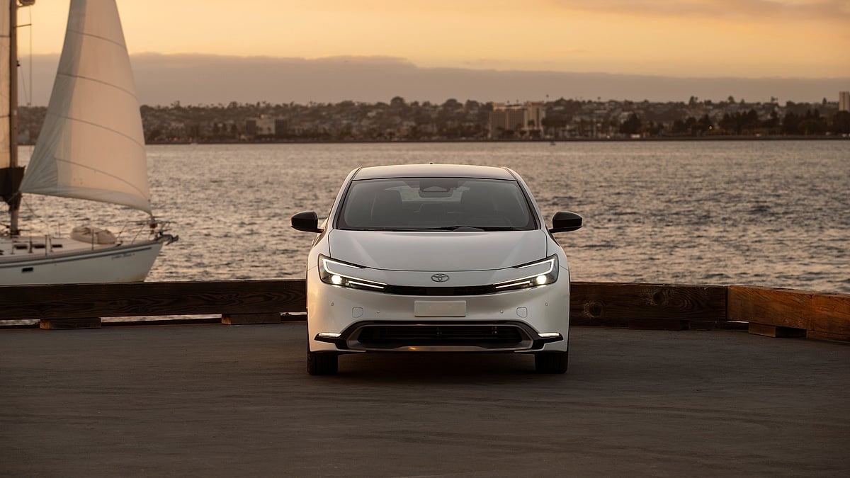 White 2024 Toyota Prius Limited shown from the front by the waterfront at sunset with a sailboat nearby.
