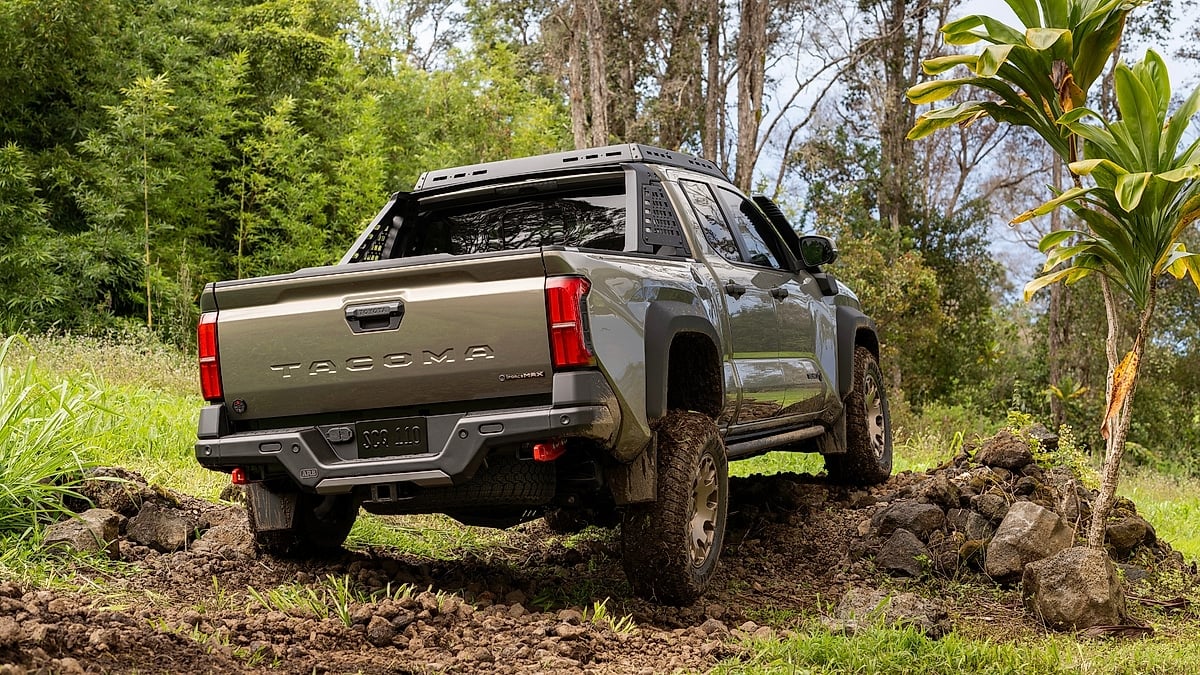 2024 Toyota Tacoma Trailhunter shown from the rear while driving over rocks on a muddy tropical trail.