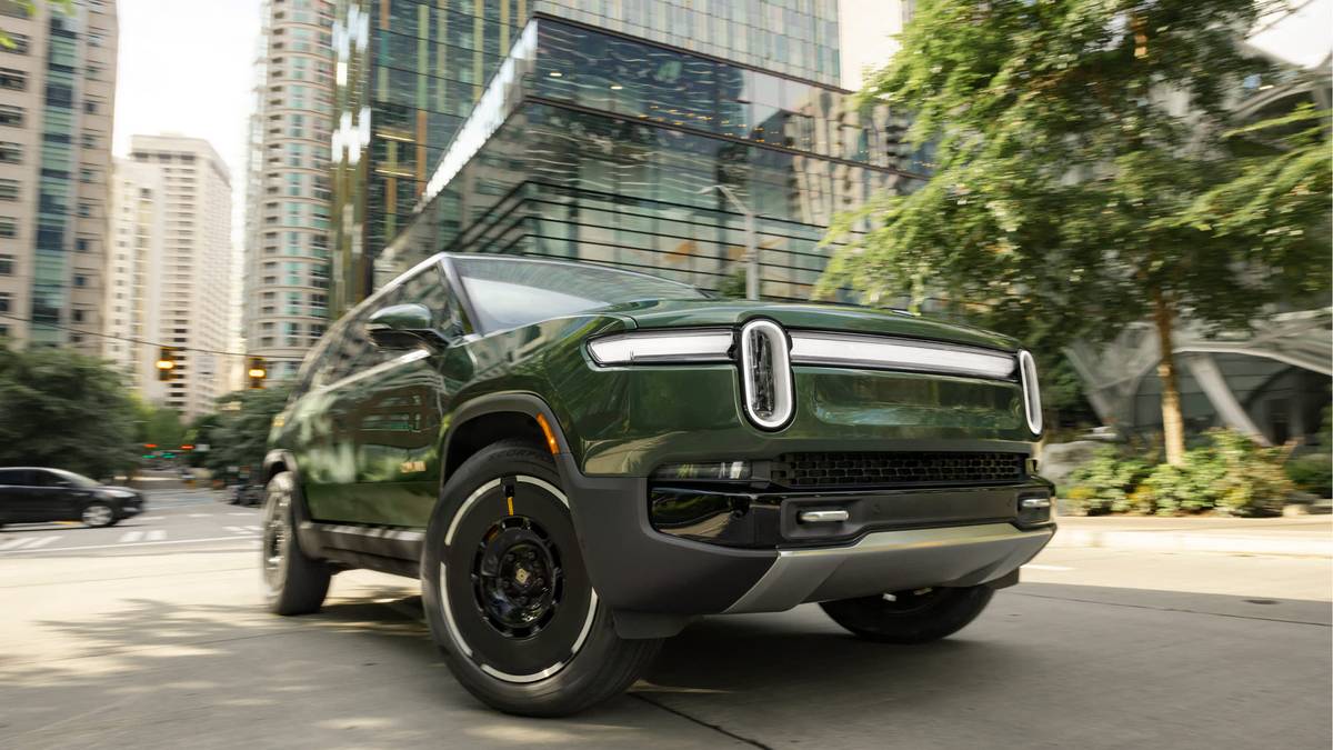 2025 Rivian R1S in Forest Green on urban street, front three-quarter view with modern glass office buildings and trees