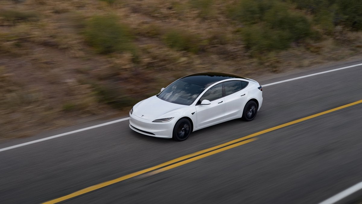 2024 Tesla Model 3 in Pearl White driving on a desert highway, front three-quarter view from elevated angle with motion-blurred scrubland background