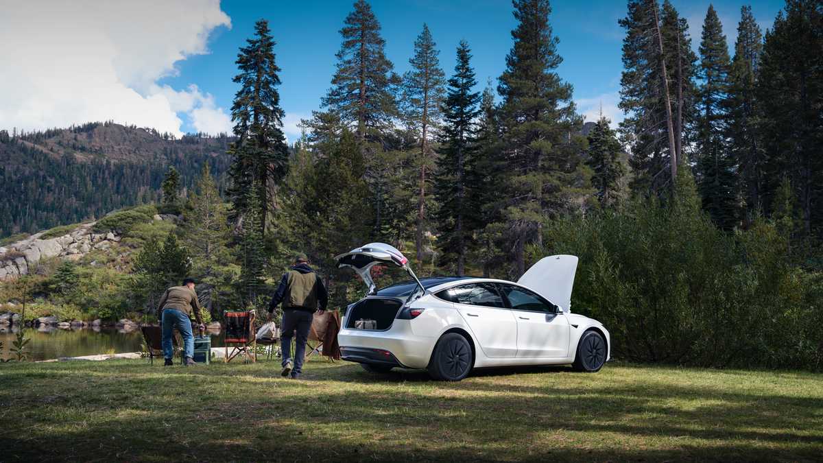 2024 Tesla Model 3 in Pearl White at a mountain campsite, rear three-quarter view with trunk open, pine forest and lake in background