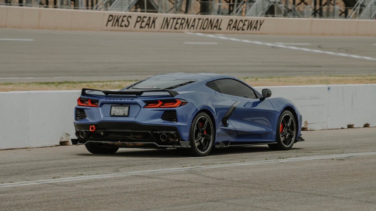 Blue 2024 Chevrolet Corvette C8 rear view on a race track, highlighting quad exhaust, rear diffuser, and mid-engine sports car proportions.