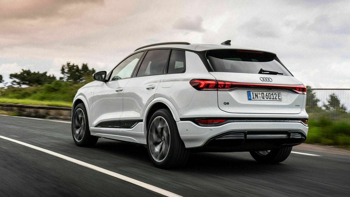 A white Audi Q6 SUV driving along a scenic road, showcasing its sleek design against a backdrop of green fields and cloudy sky.