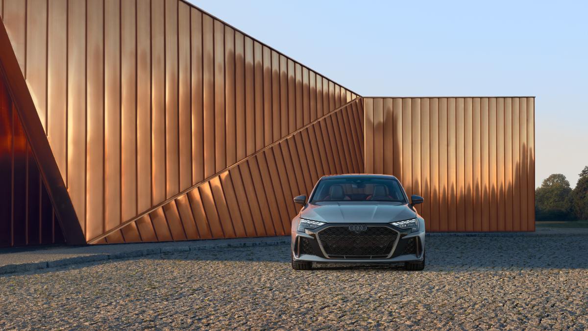 2023 Audi RS7 in metallic gray, front three-quarter view, showcasing aggressive black grille against modern copper-colored architectural backdrop at sunset.