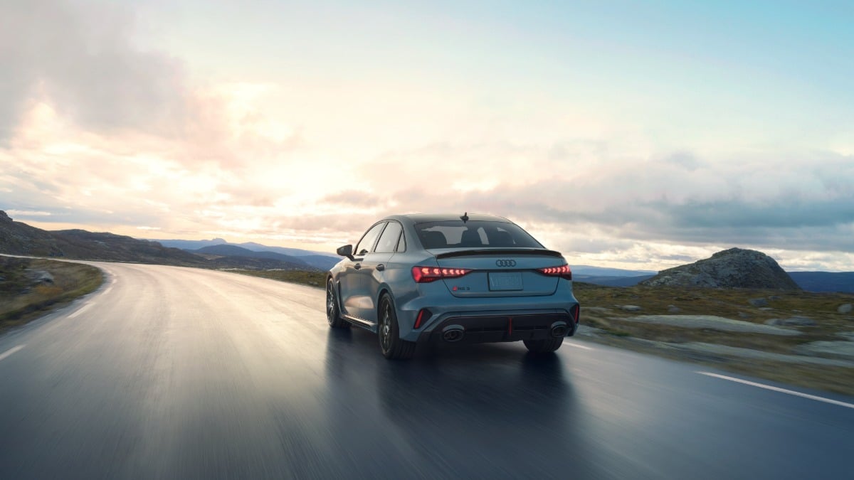 2023 Audi RS3 sedan in Nardo Gray, rear three-quarter view, driving on mountain road at sunset with LED taillights illuminated.