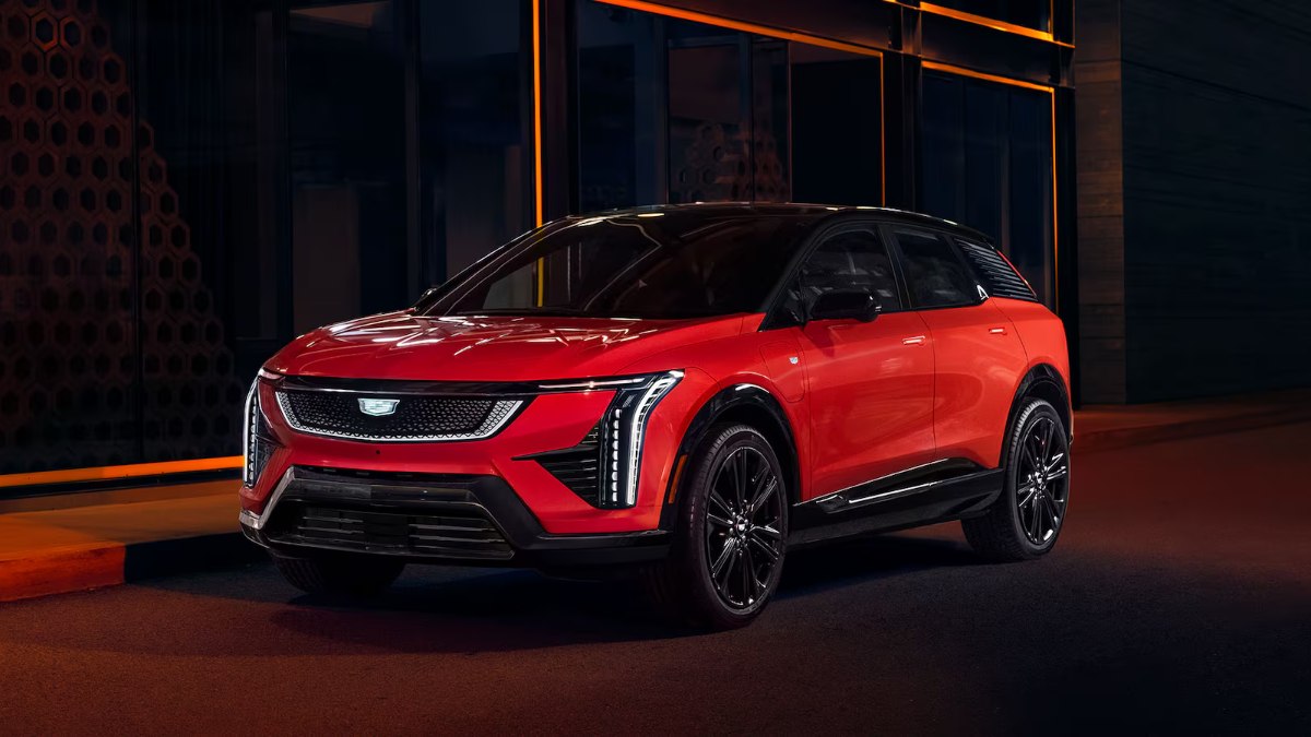 2026 Cadillac Optiq Sport in Velocity Red, front three-quarter view, featuring distinctive vertical LED lights, black-accented grille, and two-tone exterior in dramatic indoor lighting.