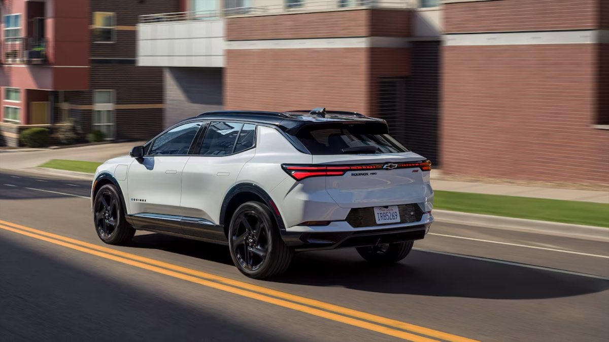 2025 Chevrolet Equinox EV in white, rear three-quarter view driving on city street with modern apartment buildings in the background.