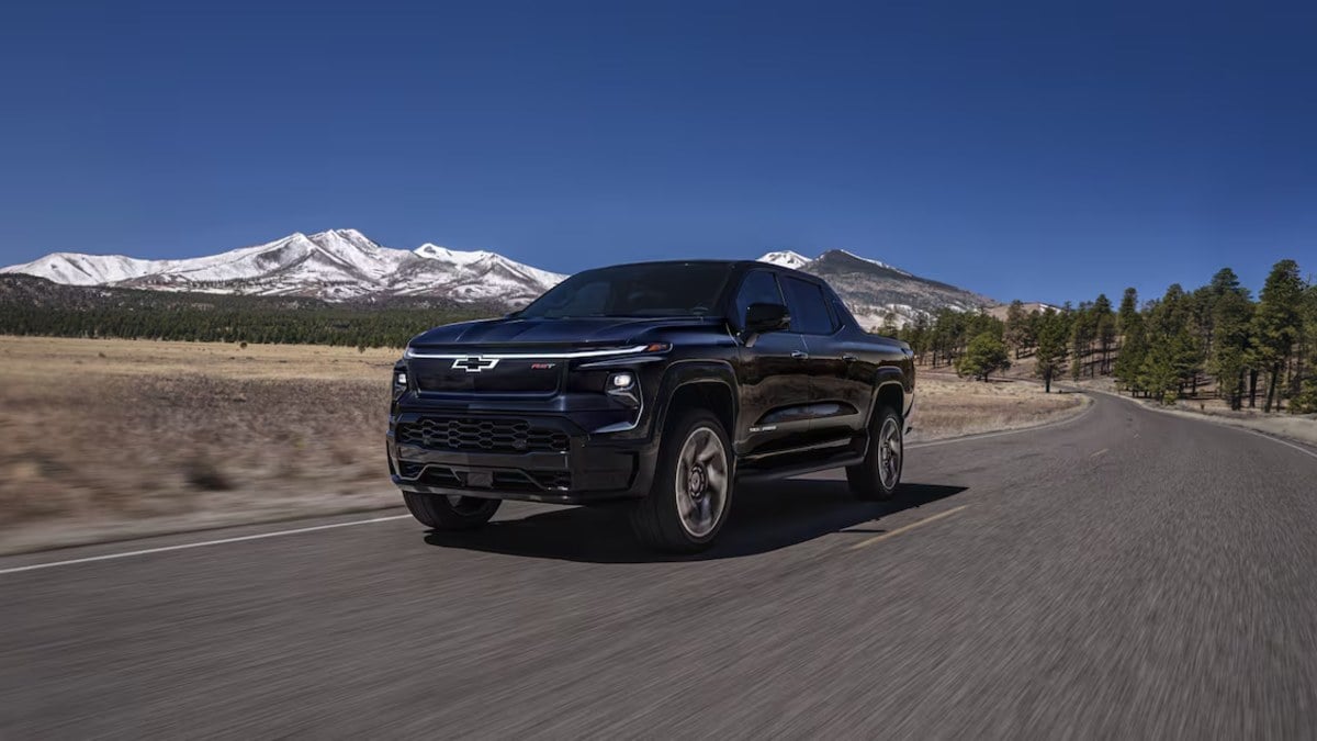 Black Chevrolet Silverado pickup truck, front three-quarter view, driving on mountain highway with snow-capped peaks and pine trees in background.