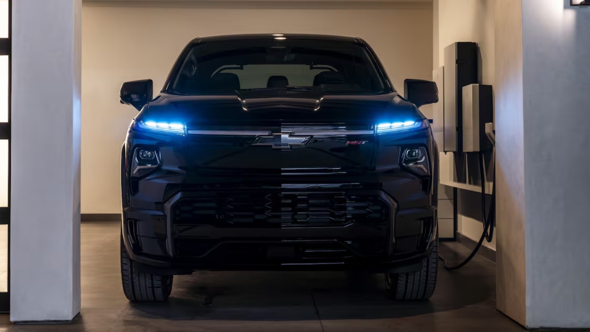 Black electric truck with illuminated headlights parked in a garage, charging station visible.