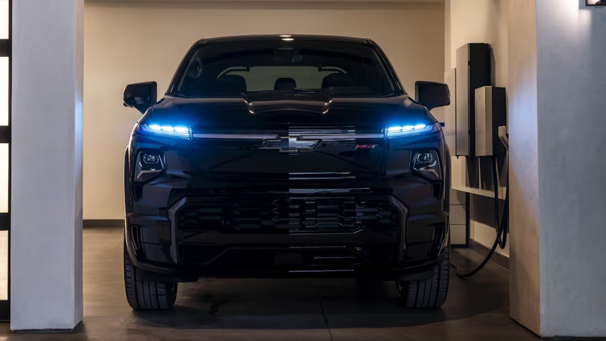 Dark blue Chevrolet Silverado EV, front view, LED headlights illuminated, connected to charging station in modern indoor garage setting.