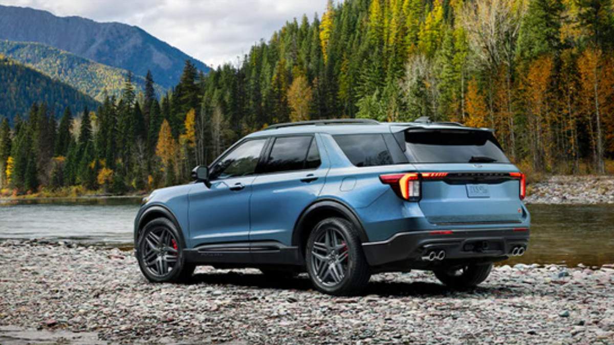 2025 Ford Explorer ST in blue, parked on rocky riverbank with forested mountain landscape behind, rear three-quarter view