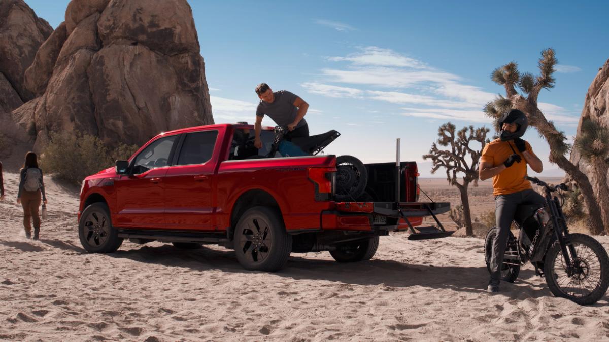 Red pickup truck in desert with people loading a motorcycle, cyclist nearby, rocky landscape.
