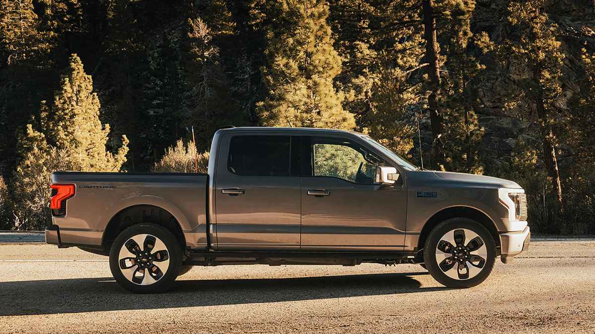 Gray pickup truck parked on a road, surrounded by tall trees in sunlight.