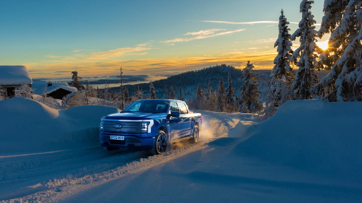 A blue truck drives through a snow-covered landscape at sunset, surrounded by tall trees and distant hills, creating a serene and adventurous mood.