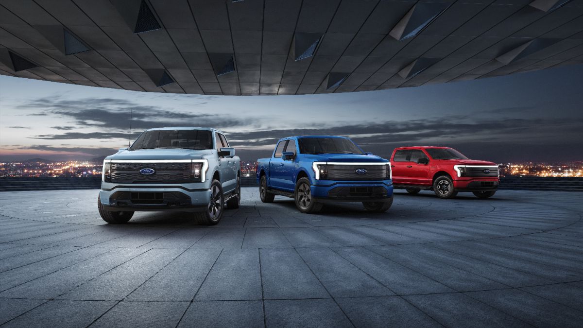 2025 Ford F-150 Lightning electric trucks in silver, blue, and red, parked under angular architectural ceiling with city lights at dusk