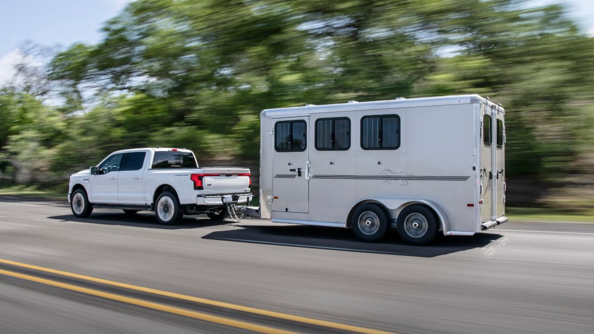 White pickup truck towing a three-horse trailer on highway with trees in background