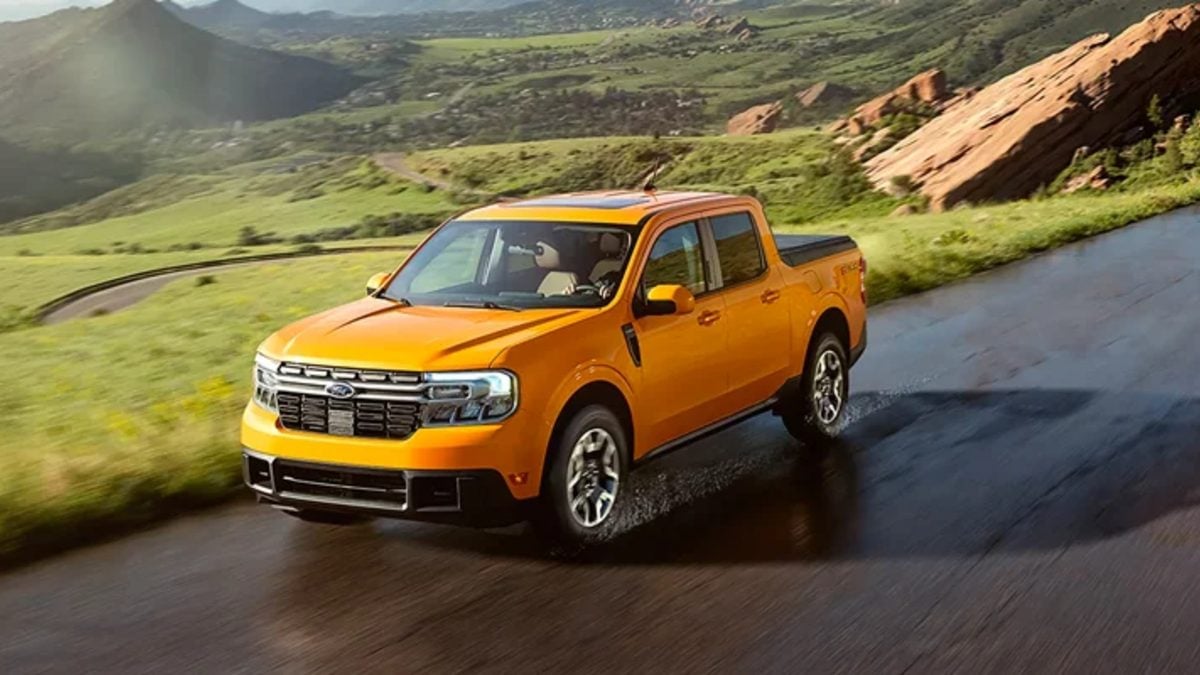 2022 Ford Maverick pickup in Cyber Orange, shown from front three-quarter view, driving on wet mountain road with green hillsides and red rock formations.