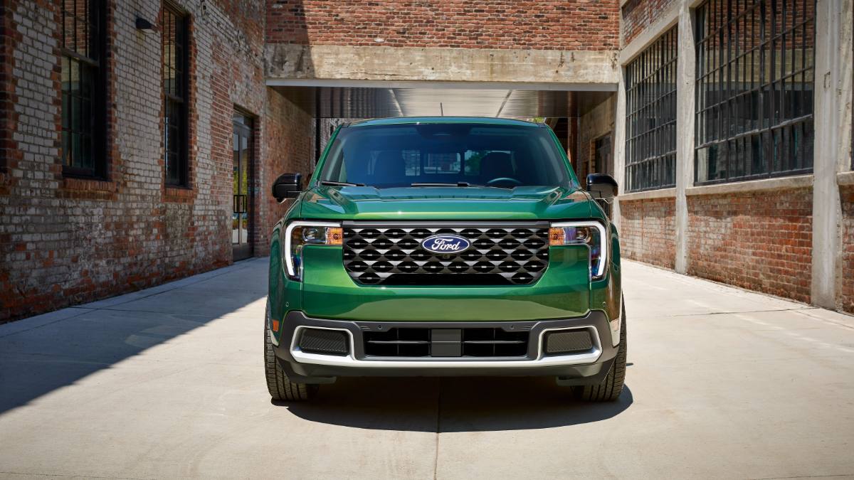 Front view of a 2023 Ford F-150 pickup truck in metallic green, showcasing its distinctive black mesh grille with chrome accents and LED lighting signature, photographed in an industrial brick building setting.