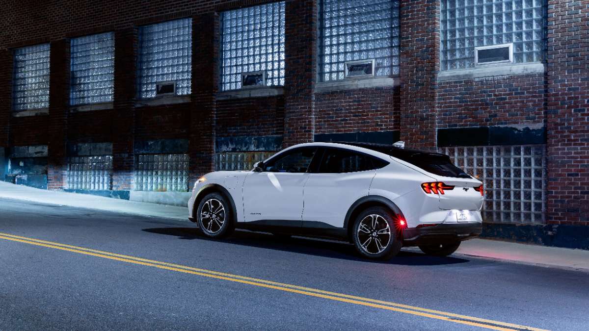 White electric SUV parked on city street, rear three-quarter view with brick building and glass block windows behind, sleek modern design with dark wheels and illuminated taillights
