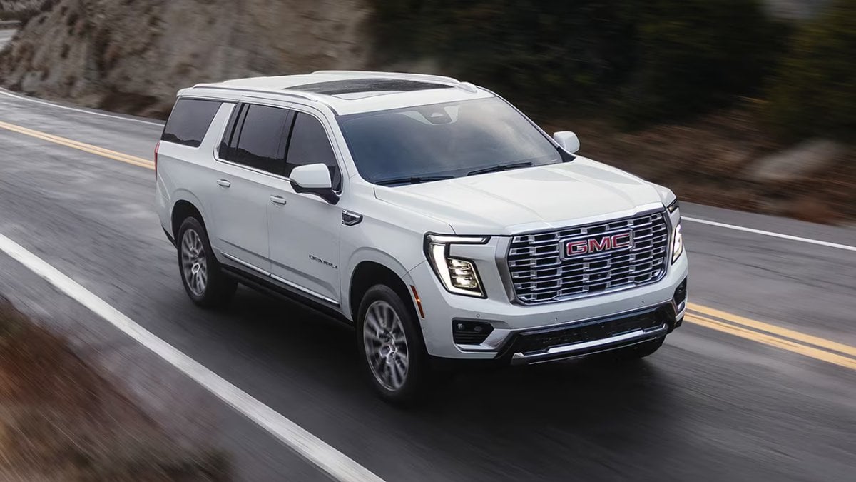 2025 GMC Yukon in pearl white, photographed from three-quarter front angle on mountain road, featuring LED headlights, chrome grille, and panoramic sunroof.