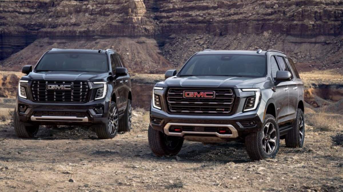 Two 2024 GMC Yukon AT4 SUVs in metallic gray, photographed at three-quarter front angle against rugged desert cliffs, featuring distinctive LED lighting and off-road trim.