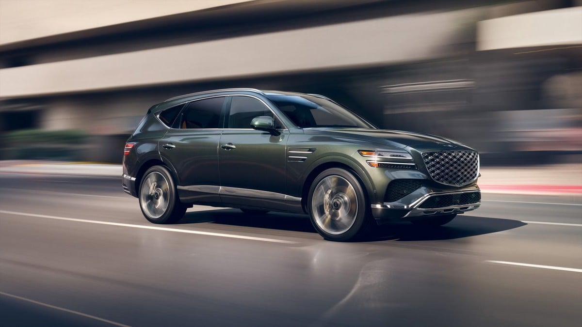 Dark green 2025 Genesis GV80 SUV in motion, shot from side profile on blurred urban roadway with dynamic wheel and background blur