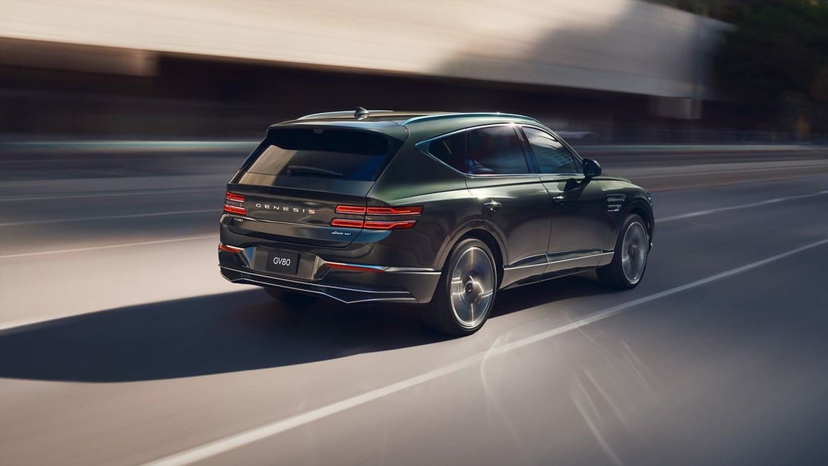 Dark green 2025 Genesis GV80 SUV in motion, rear three-quarter view on highway with blurred background and dynamic wheel spin