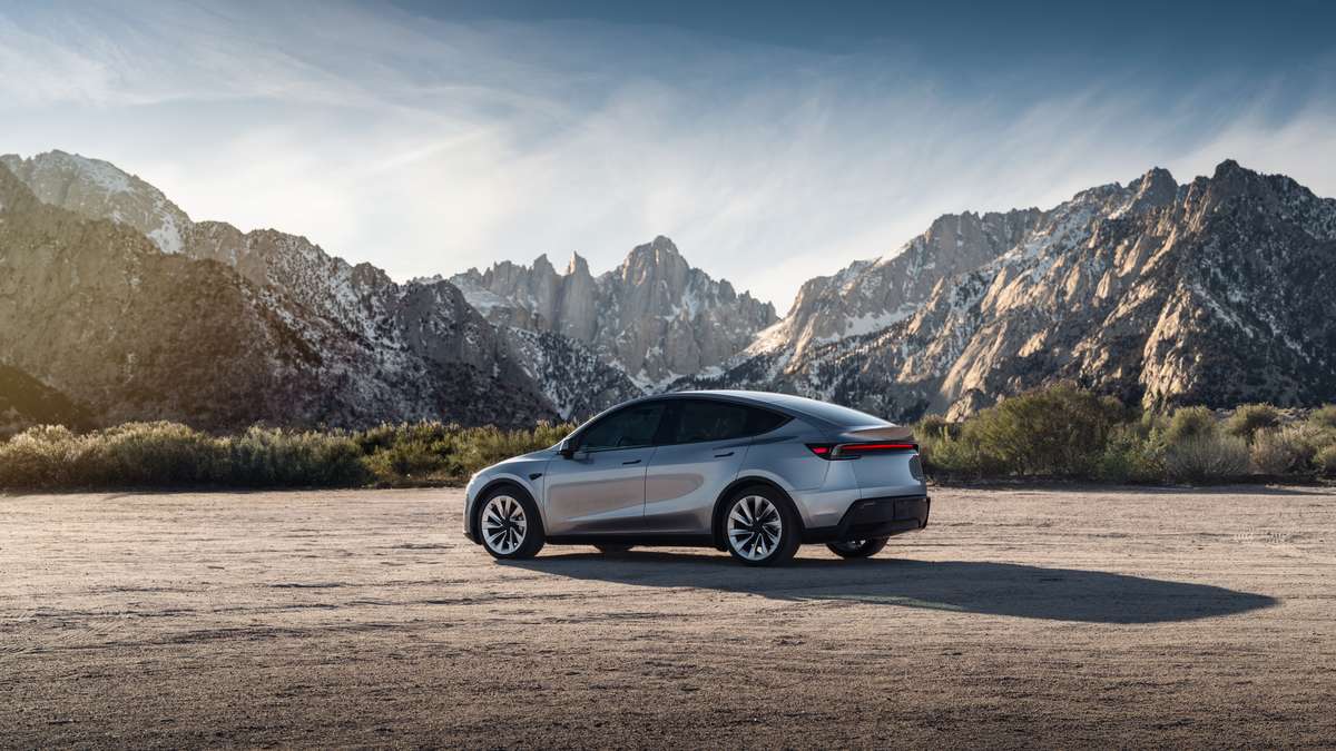 A sleek, silver electric car driving on a winding road surrounded by mountains, illuminated by bright sunlight.