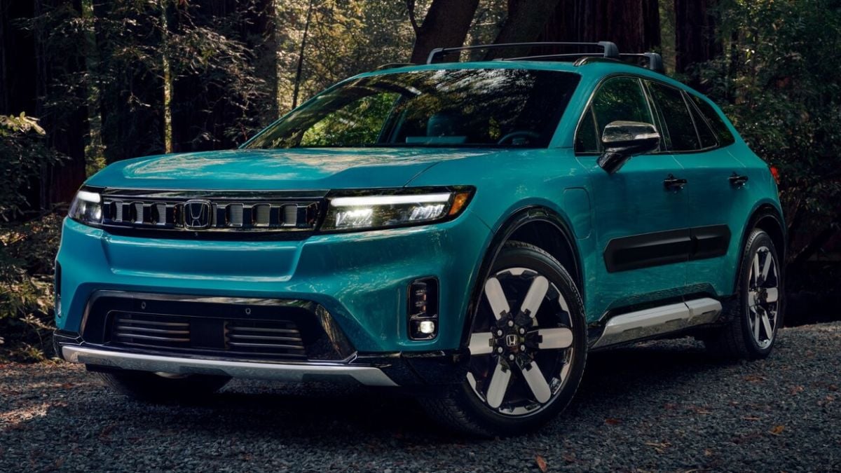 A teal Honda Prologue with a modern design is parked on a gravel path in a forest setting. Sunlight filters through the trees, highlighting the vehicle's curves.