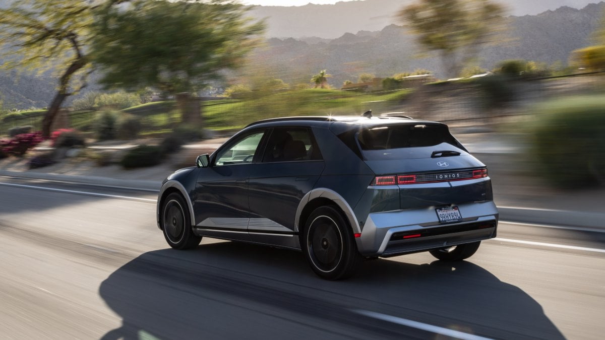 A dark-colored car driving on a road with blurred trees and mountains in the background.