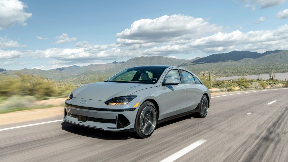 2024 Hyundai Ioniq 6 in silver, front three-quarter view, driving on desert highway with mountains and saguaro cacti in background. Sleek aerodynamic design with distinctive LED lighting.
