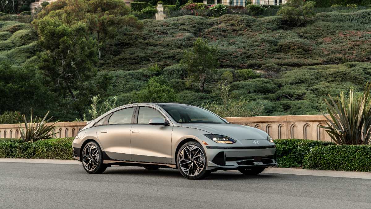 Silver, modern electric car parked on a road beside lush greenery and a stone railing, conveying an upscale, eco-friendly vibe under a clear sky.