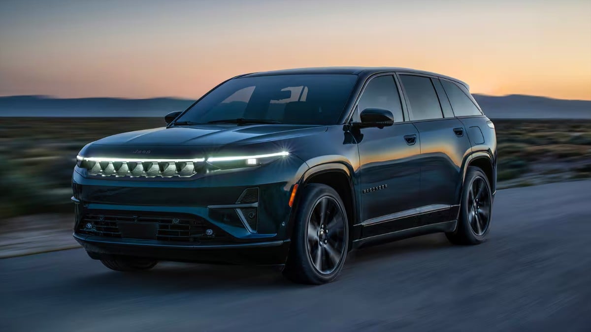 Dark blue 2025 Jeep Wagoneer S electric SUV driving on highway at sunset with distinctive LED headlight design