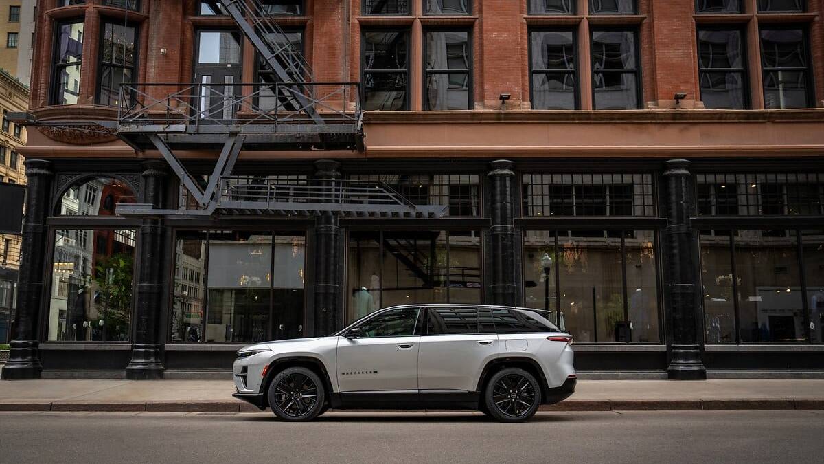 2025 Jeep Wagoneer S EV in white, parked beside urban brick building with large glass storefront, side profile showing sleek electric SUV design