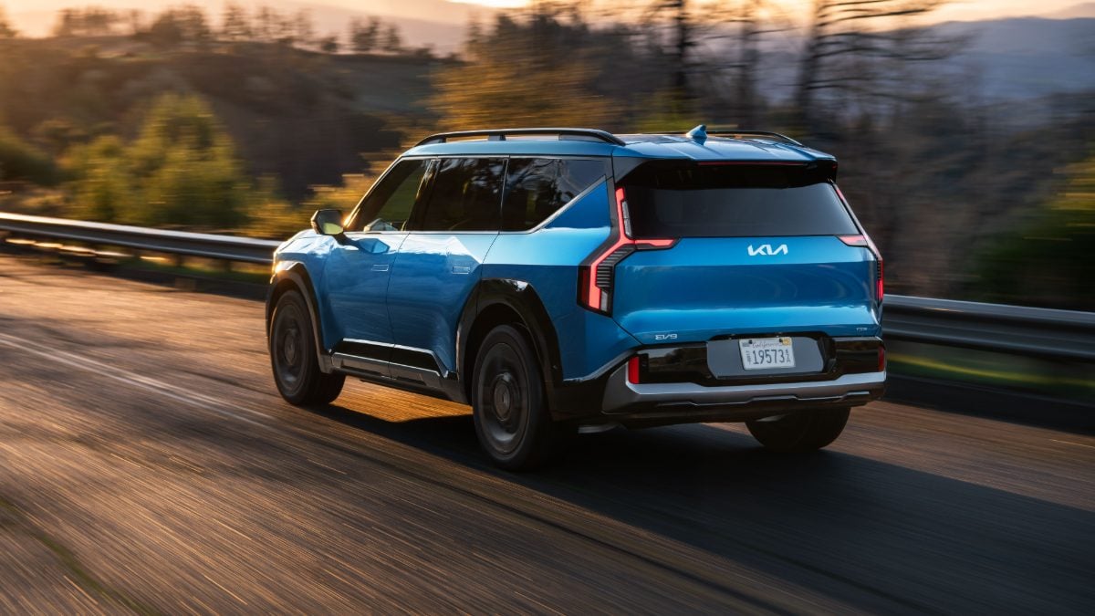 Blue 2025 Kia EV9 electric SUV driving on a scenic road at sunset, rear three-quarter view.