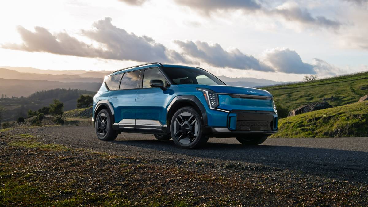 Blue 2025 Kia EV9 EV SUV parked on a mountain road with dramatic clouds, front three-quarter angle.