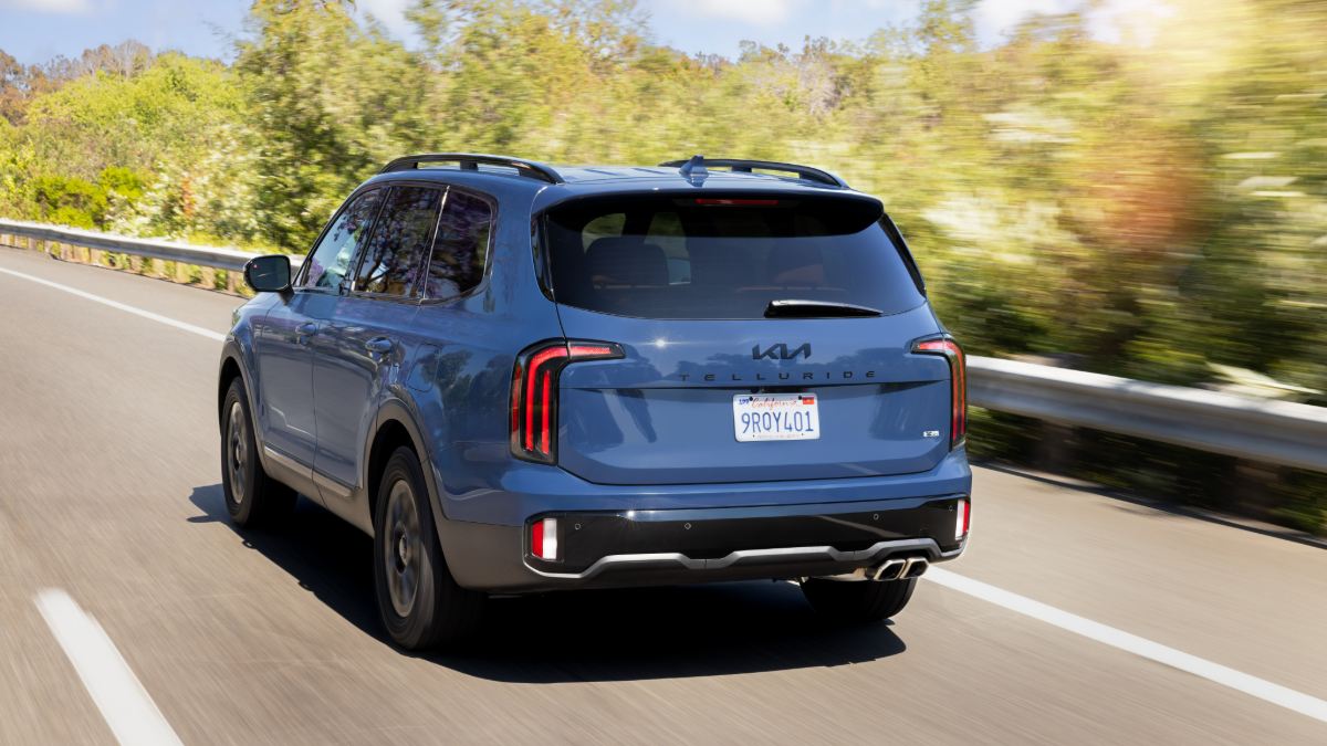 A blue 2025 Kia Telluride SUV shown from the rear three-quarter view, driving on a curved road with blurred foliage in the background.
