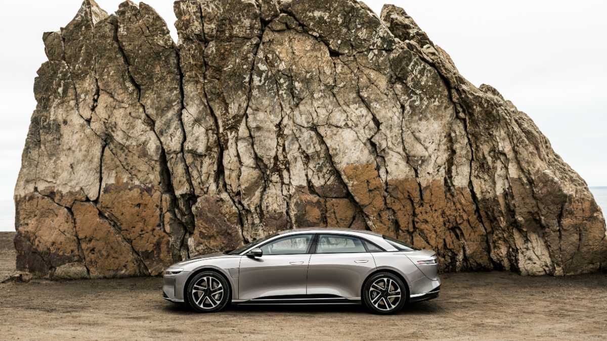 A silver Lucid Air luxury electric sedan is shown in side profile view against a dramatic rocky cliff backdrop, highlighting the car's sleek aerodynamic design and distinctive silhouette.