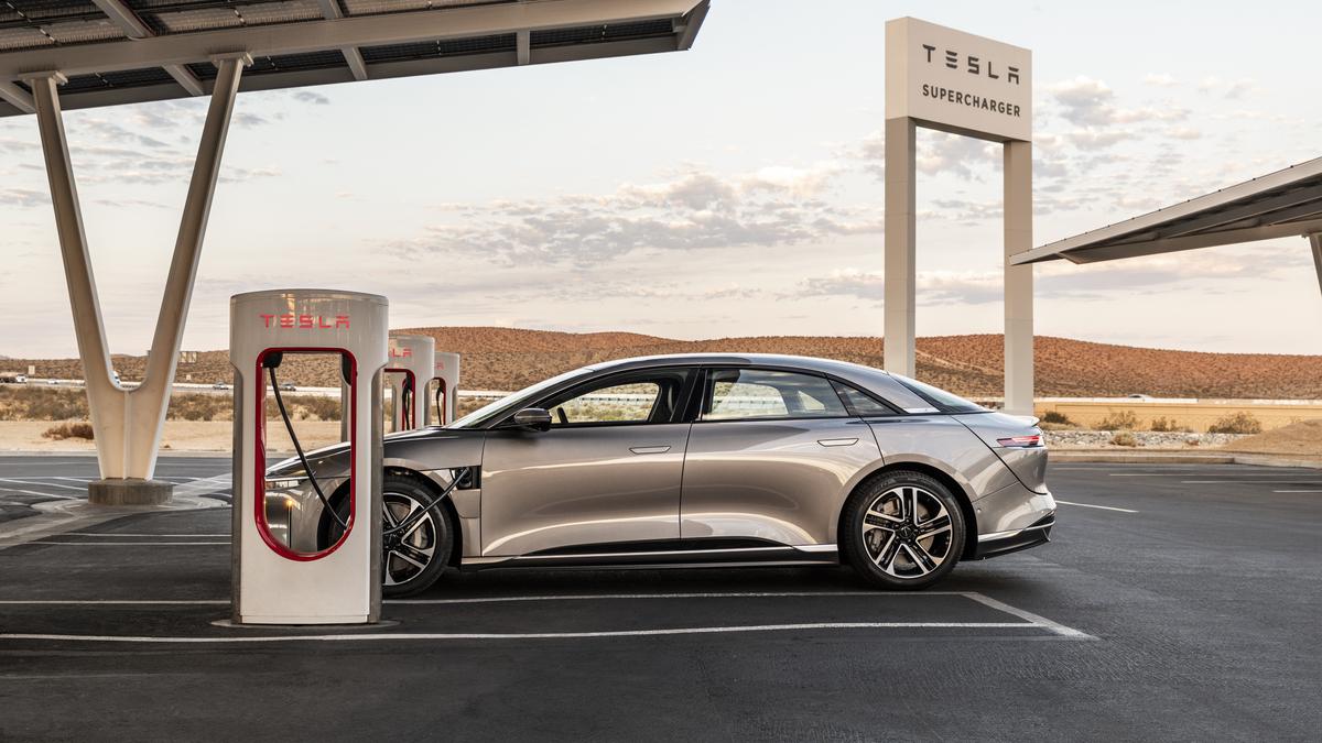 Image of a 2025 Lucid Air electric vehicle in a gray color, photographed from the front three-quarter angle while charging. The vehicle's front grille, headlights, and charging port are clearly visible.