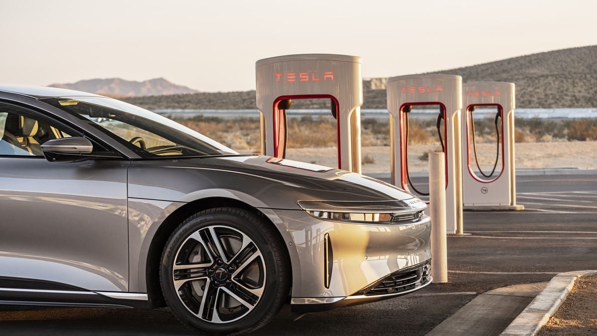 A 2025 Lucid Air parked at Tesla charging stations in a desert landscape, with mountains in the background under a clear sky.