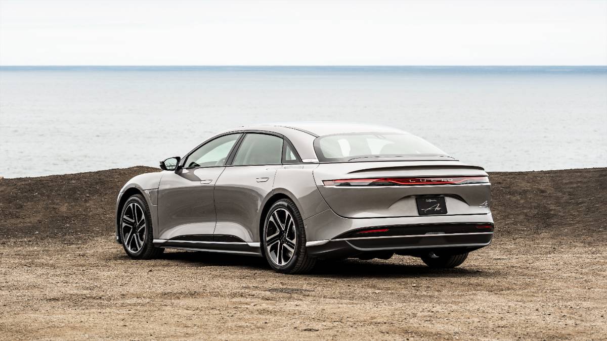 Silver 2025 Lucid Air electric sedan, rear three-quarter view on rocky coastal terrain with ocean horizon