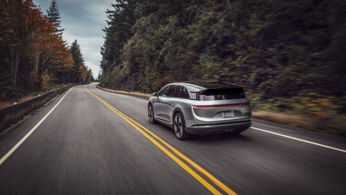 Silver Lucid Air electric luxury sedan shown from rear three-quarter view, driving on curved mountain road surrounded by autumn forest.