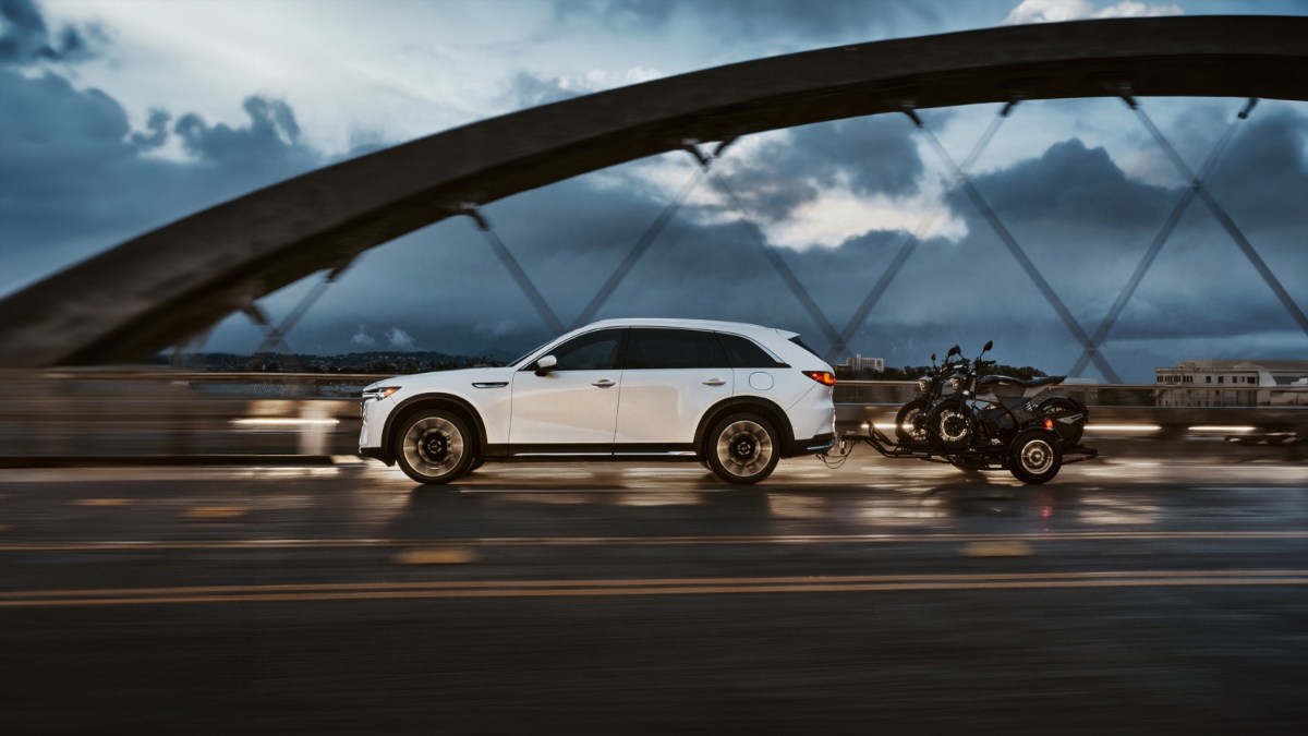 White 2025 Mazda CX90 SUV driving on wet road with motorcycle, dramatic cloudy bridge background