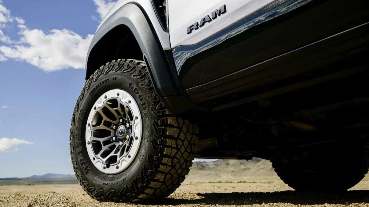 Close-up detail shot of a Ram truck's off-road wheel and tire combination, featuring an aggressive all-terrain tire mounted on a two-tone aluminum wheel, set against a desert landscape background.
