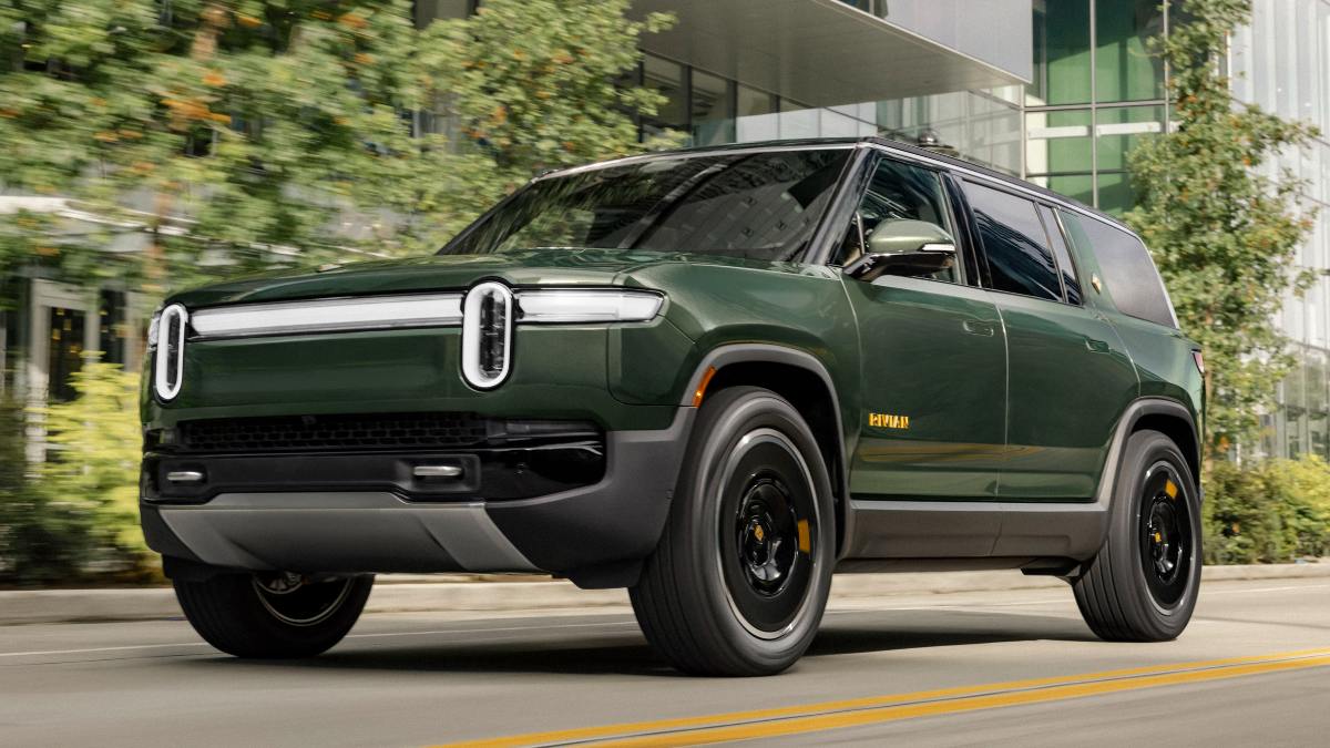 A dark green 2024 Rivian R1S electric SUV shown from a front three-quarter angle, featuring distinctive vertical LED headlights and black wheel accents against an urban background with trees.