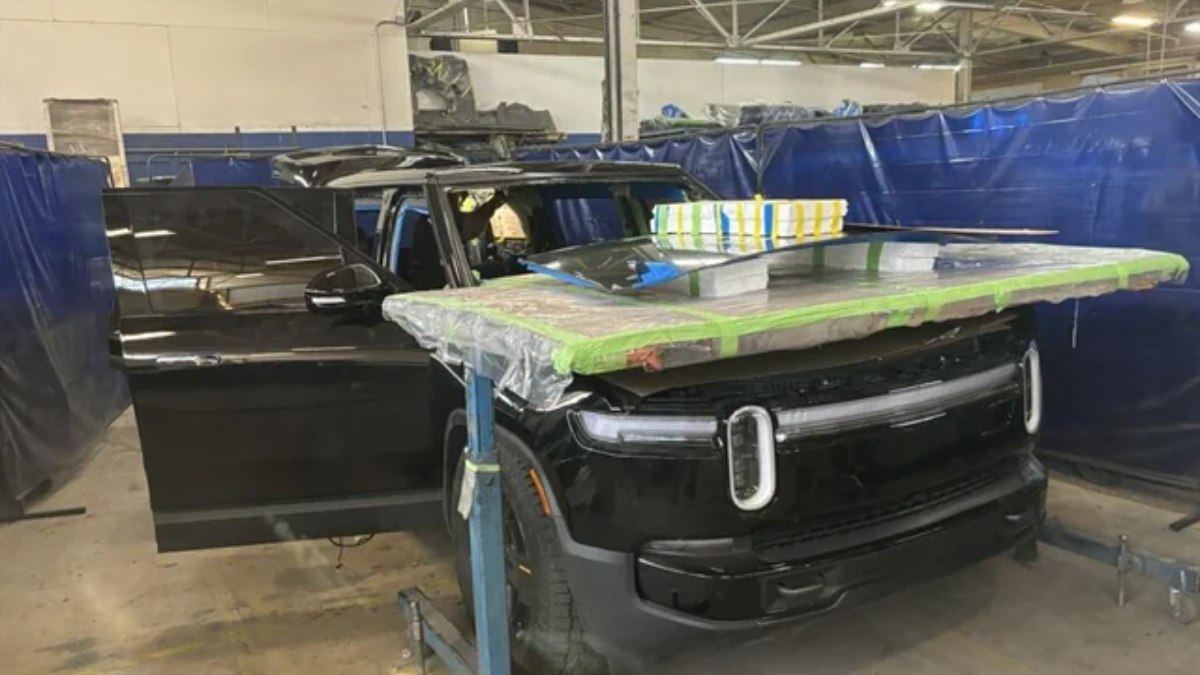 A black vehicle is parked in a workshop, covered with plastic and tape, with a large sheet resting on its roof.