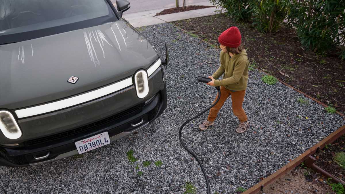 A person in a red hat connects an electric cable to a green truck on a gravel driveway, conveying a sense of eco-friendly technology and sustainability.