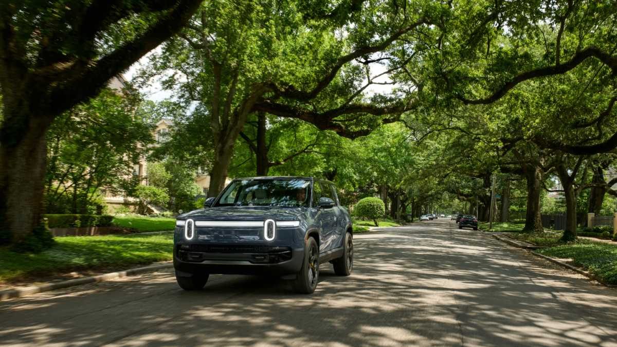 Dark gray Rivian electric SUV driving on tree-lined residential street with dappled sunlight