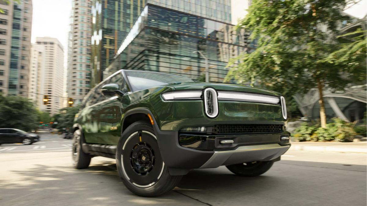 Dark green Rivian R1S electric SUV driving on city street with glass buildings behind